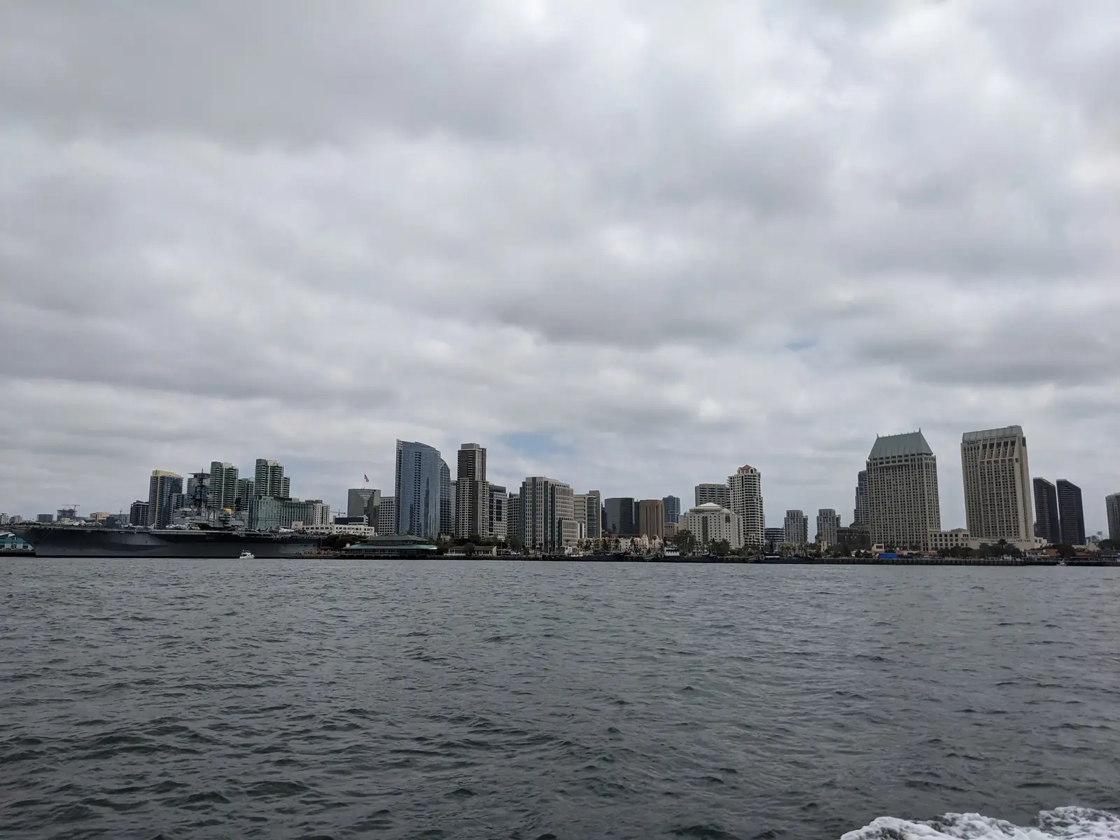 The downtown San Diego skyline viewed across the bay under an overcast sky, with the USS Midway Museum visible at the waterline.