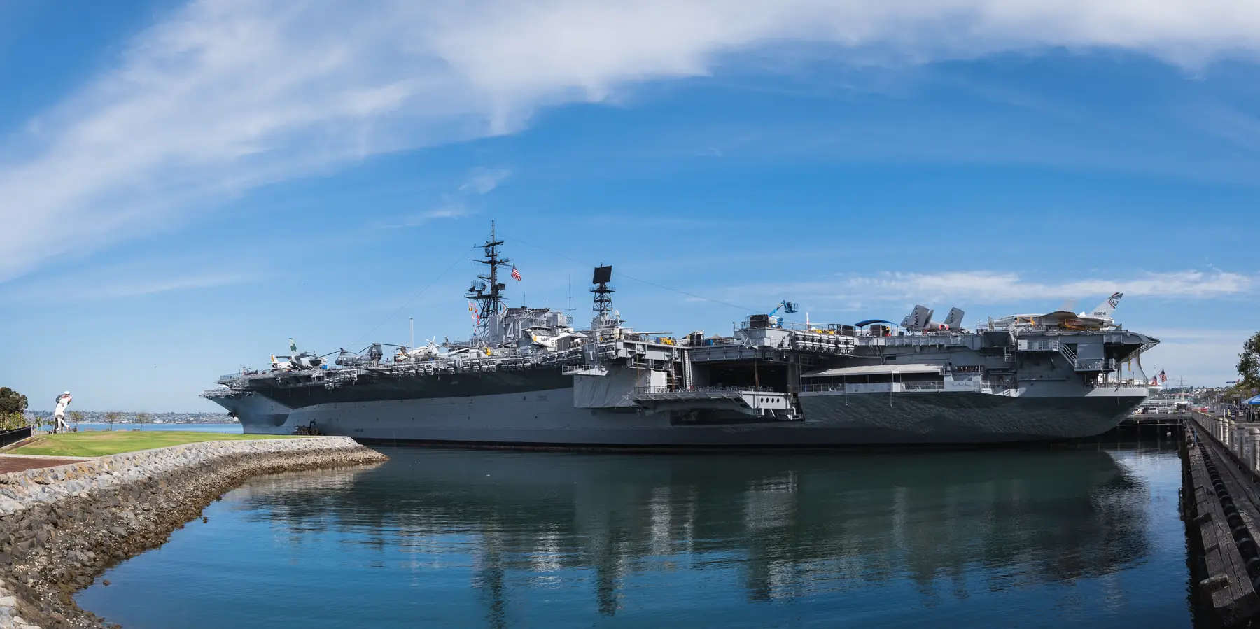 A wide view of the USS Midway Museum, a retired aircraft carrier docked at the Embarcadero, under a blue sky with light clouds.