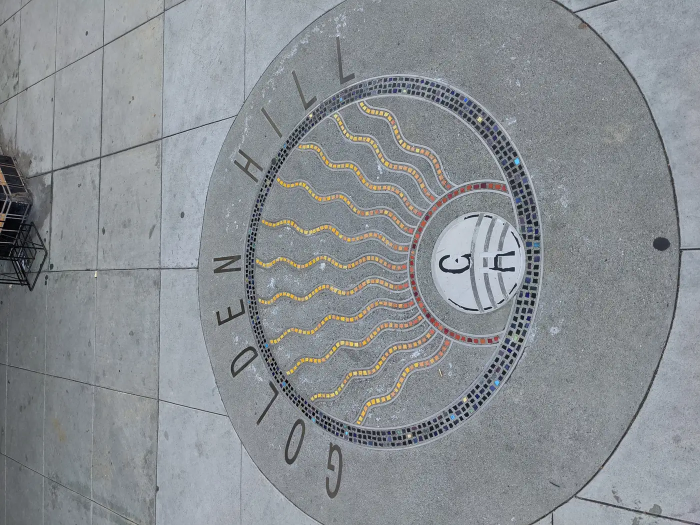 A circular sidewalk medallion reading Golden Hill with a mosaic sunburst pattern in orange, yellow, and blue tile.
