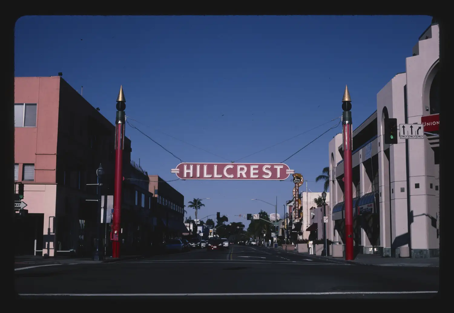 A vintage photograph of the red neon Hillcrest sign arching over University Avenue between two spired posts, flanked by storefronts.