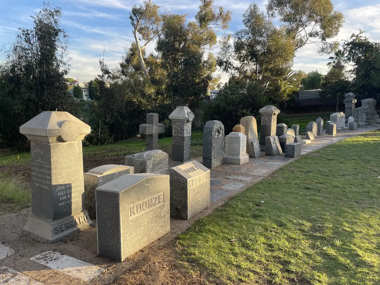 Weathered granite headstones from the late 1800s stand in rows at Calvary Pioneer Memorial Park in Mission Hills, backed by eucalyptus trees.