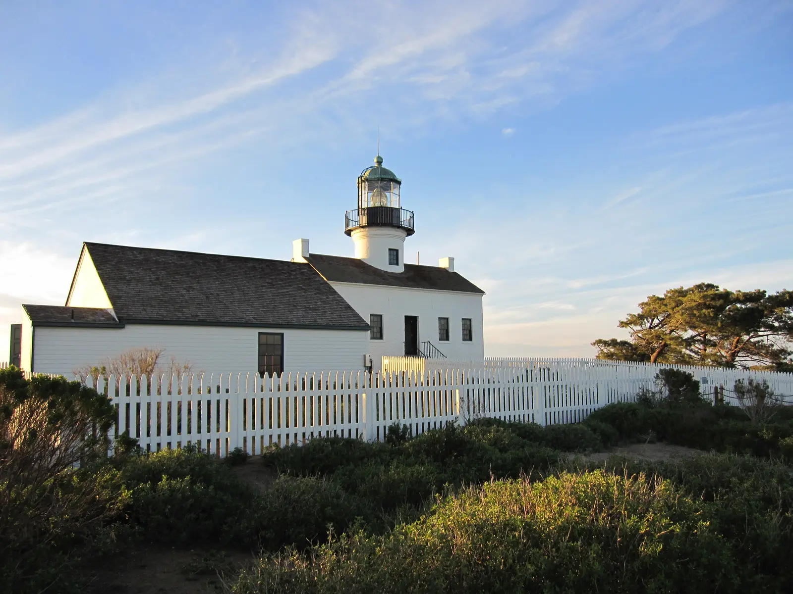 The Old Point Loma Lighthouse at golden hour, a white two-story building with a green-topped lantern room, behind a white picket fence.