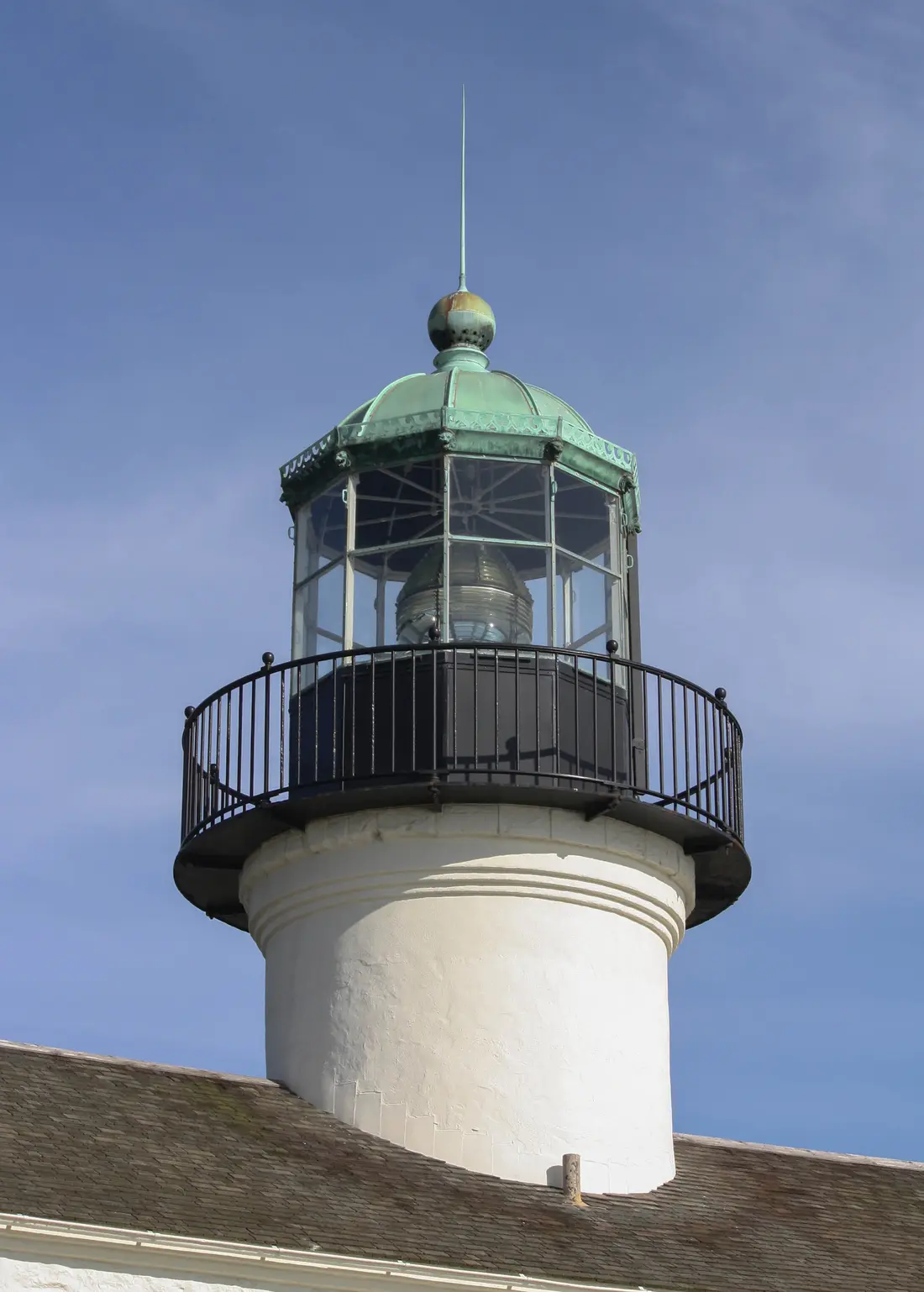 A close-up of the lighthouse's lantern room, showing its green copper dome, glass panes, and iron railing against a blue sky.
